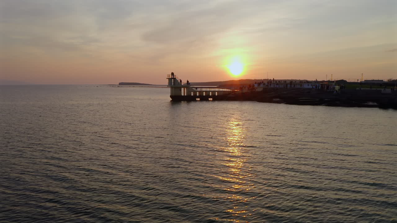 Shimmering water surrounds diving tower at sunset as soft clouds glow in orange hues, drone descends
