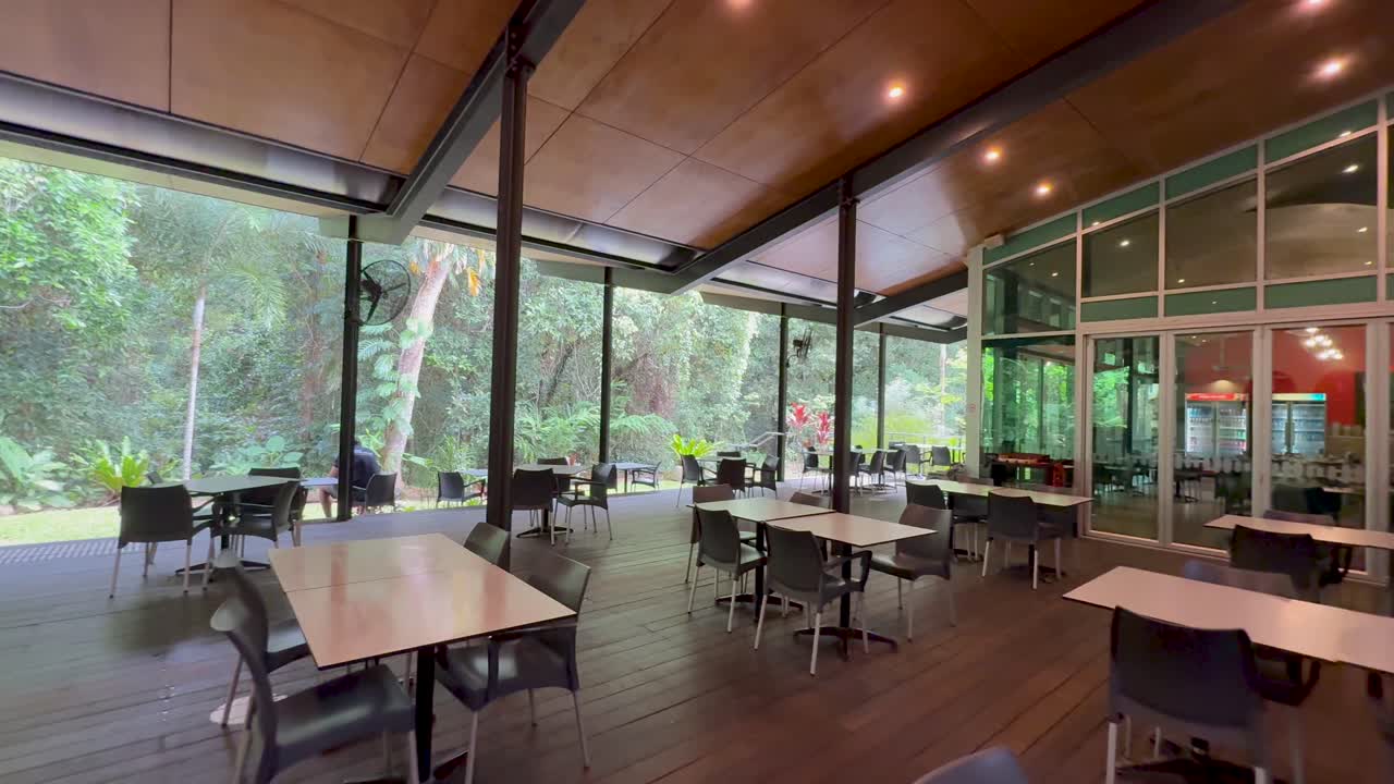 Wide-angle pan of modern rainforest cafe, woman walking, natural light, glass walls, lush greenery