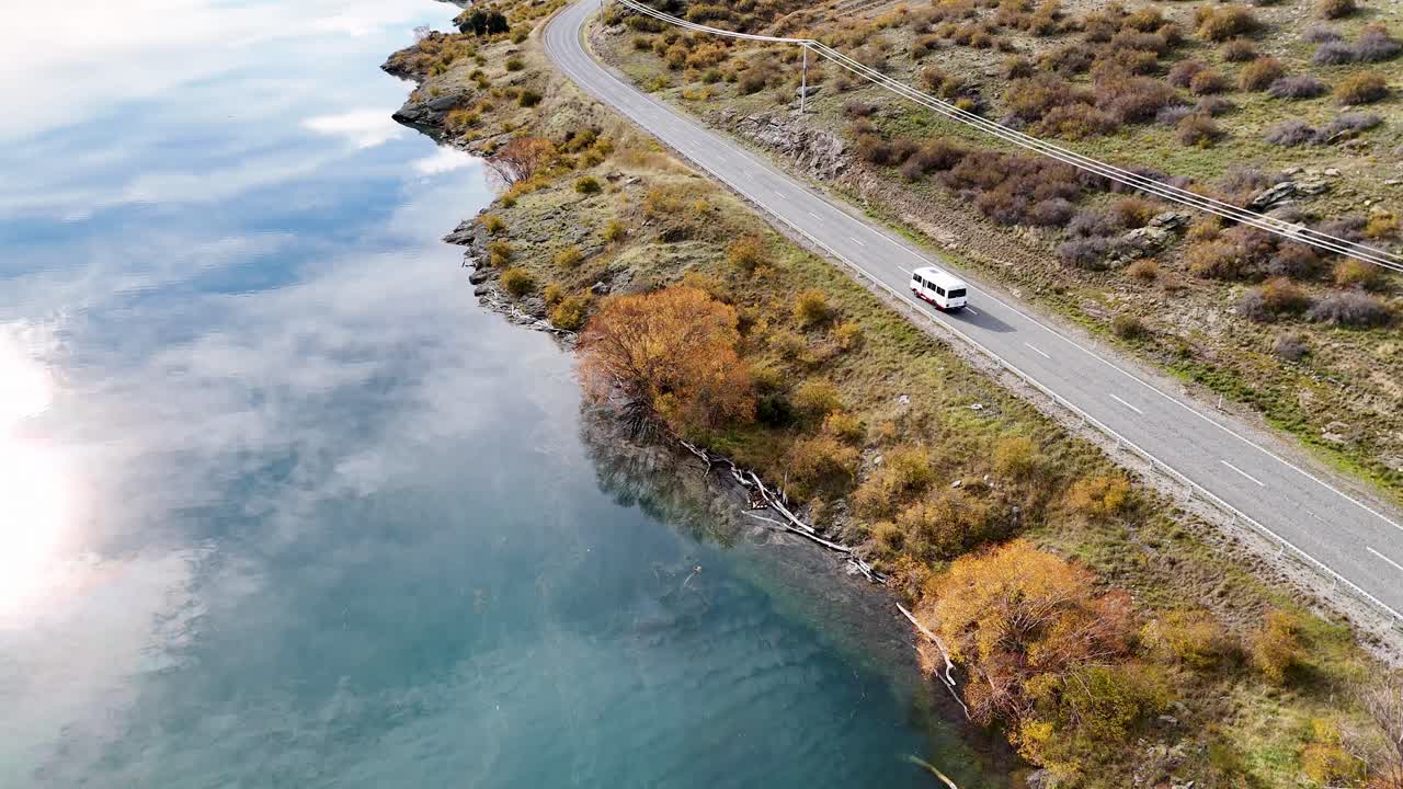 Drone captures a serene lakeside road in Cromwell, New Zealand, with autumn foliage and calm waters reflecting the sky