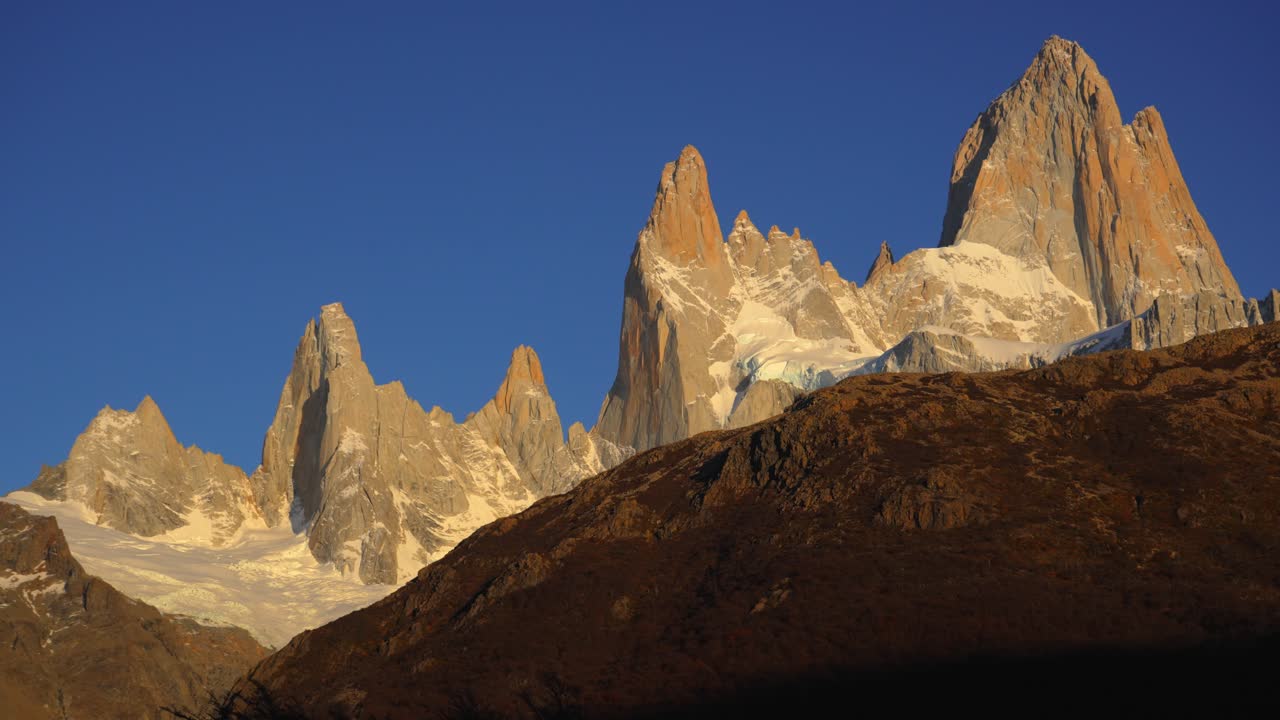 Catena montuosa del Fitz Roy bagnata da luce dorata contro un cielo azzurro limpido durante l'alba, ripresa panoramica.