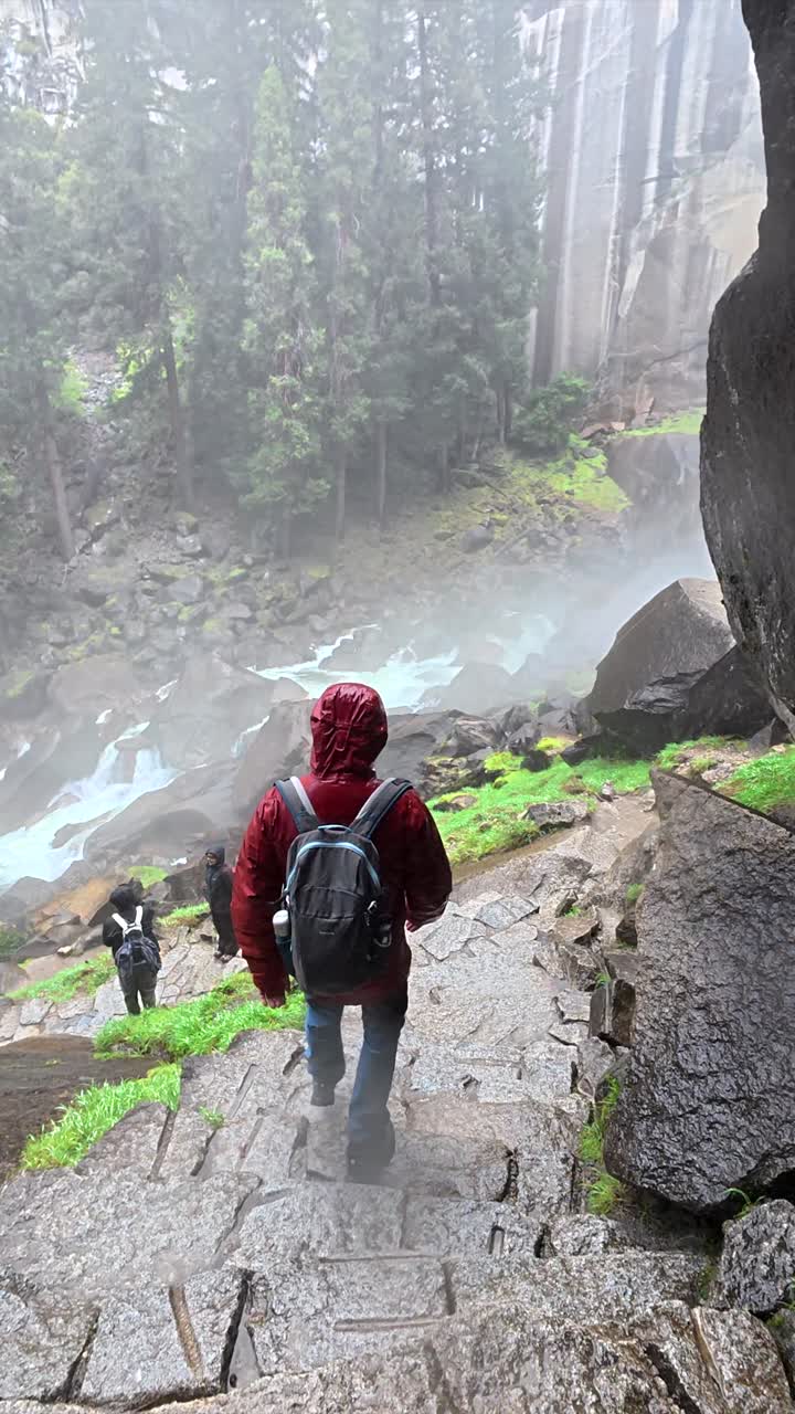 Caminhante desce por um caminho de pedra molhado em direção a uma cachoeira majestosa.