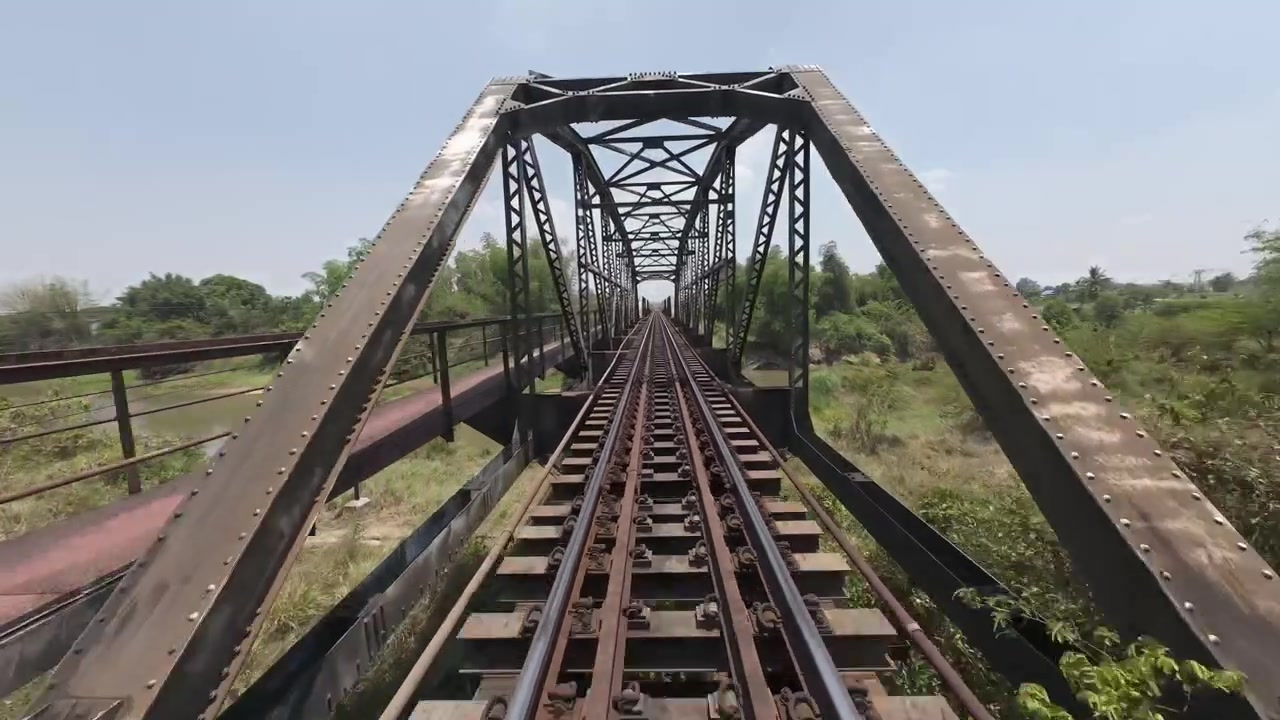 Vuelo FPV a baja altura y alta velocidad a lo largo de vías de ferrocarril cruzando un puente metálico histórico a través de exuberantes paisajes tailandeses.