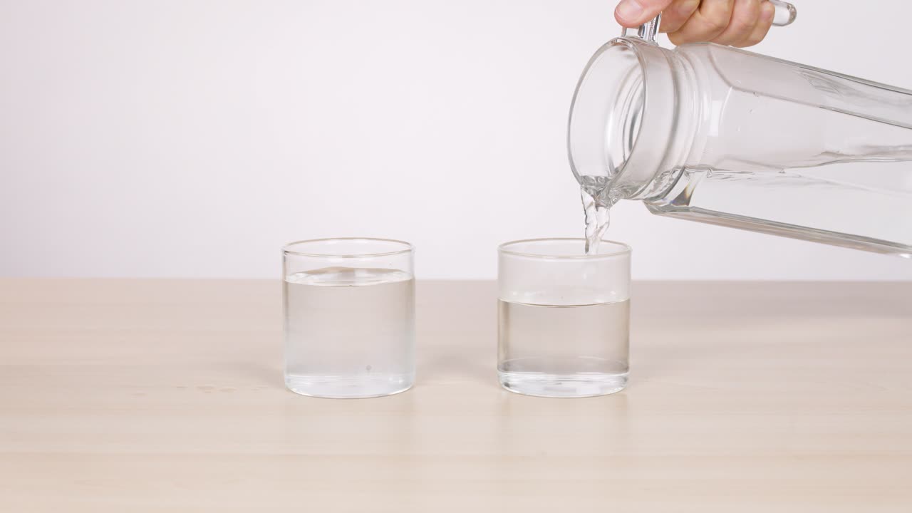 Hand pours water from glass pitcher into two glasses on table, bright even lighting