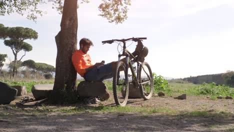 ciclista con bicicleta sentado en una roca bajo un árbol tomando selfies frente al antiguo acueducto romano. joven atractivo hombre atlético con ropa deportiva naranja y mochila en el parco degli acquedotti en roma