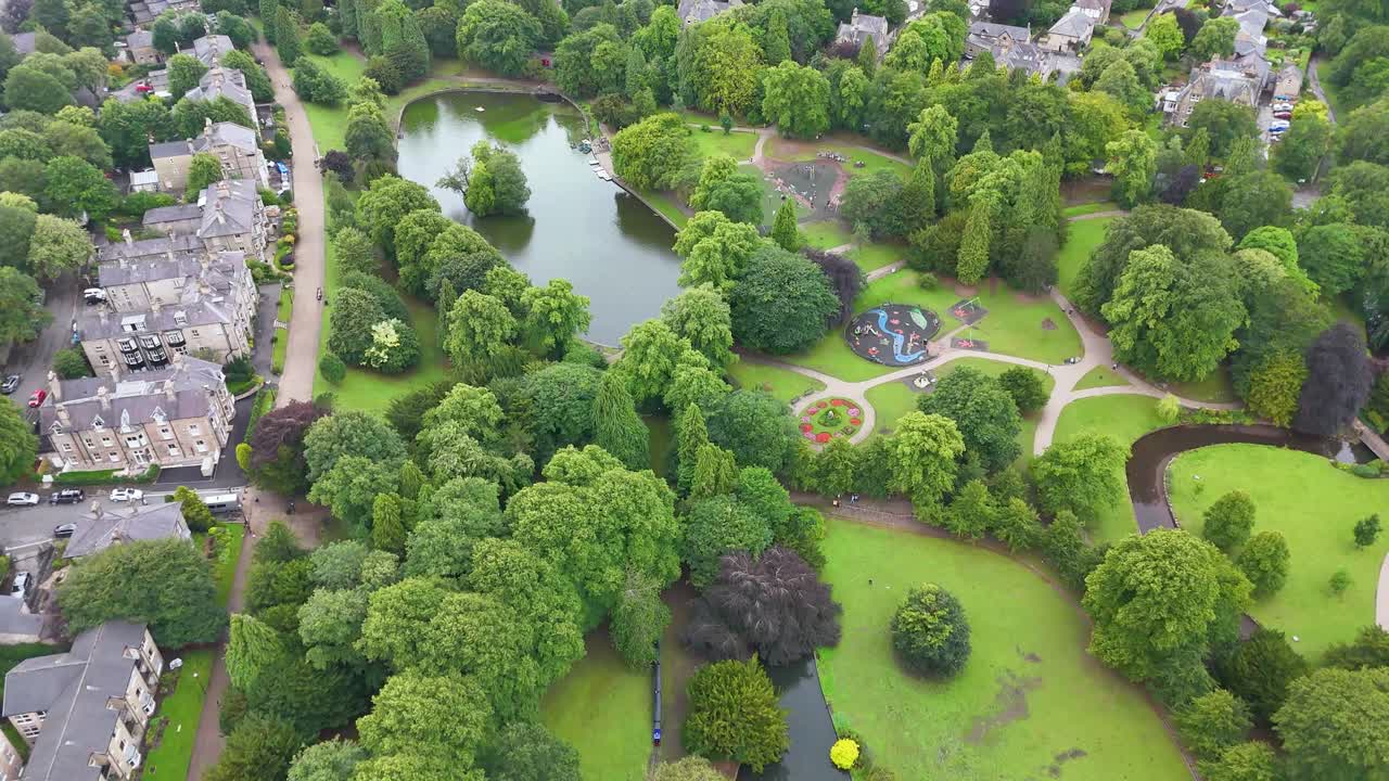 Drone footage glides above a lush green park with ponds, walking paths, and adjacent residential streets in Buxton, Derbyshire, under soft daylight