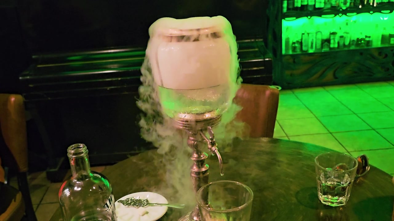 Lähikuva dramaattisesta absinttitarjoilusta, jossa louche-suihkulähde kaataa lasiin, samalla kun sumu leviää pyöreän baaripöydän, viinapullon ja lusikan yli, ja himmeä vihreä cocktail-loungen valaistus yöllä.