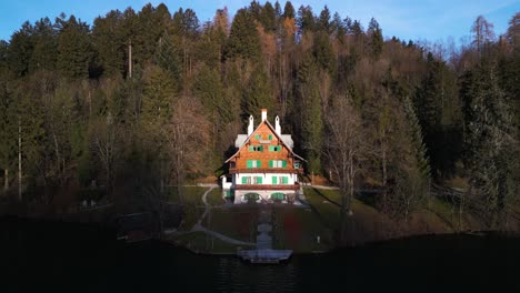 drone pullback away from sunlit wooden cabin in between leafless trees in forest by lake