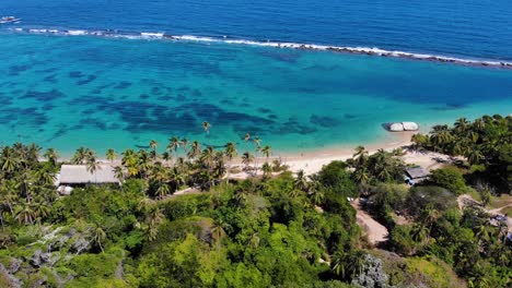 Aerial-view-away-from-a-white-sand-beach,-palm-trees-and-shallow,-blue-ocean,-in-Tayrona-national-natural-park,-sunny-day,-in-Caribbean-Colombia,-South-America---tilt-up,-drone-shot