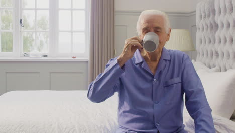 portrait of senior man at home wearing pyjamas sitting on bed having morning cup of tea