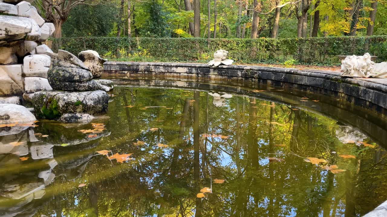 filming of a fountain in the Jardin del Principe where the reflection appears in the water of the trees, and we see sculpted heads in the fountain where a stream of water will come out of them