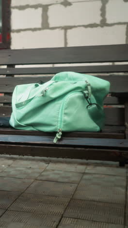 bolsa de color menta y paraguas azules descansando en un banco oscilante de madera afuera, con el fondo que muestra un edificio parcialmente construido, vegetación y pavimento entrelazado