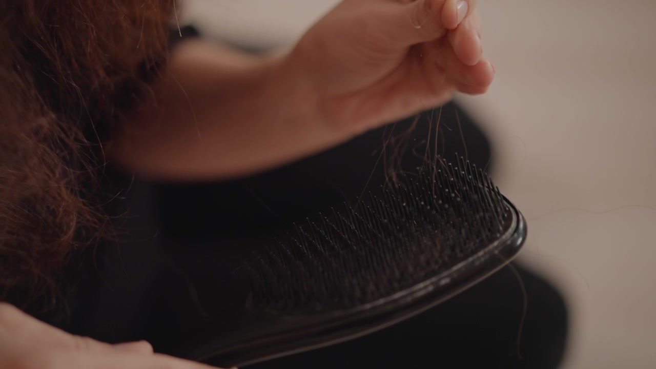 Adult hand removes trapped hair from dark cushion hairbrush, bending dense short bristles under warm indoor light, with careful grooming, tactile movement, and detailed close focus