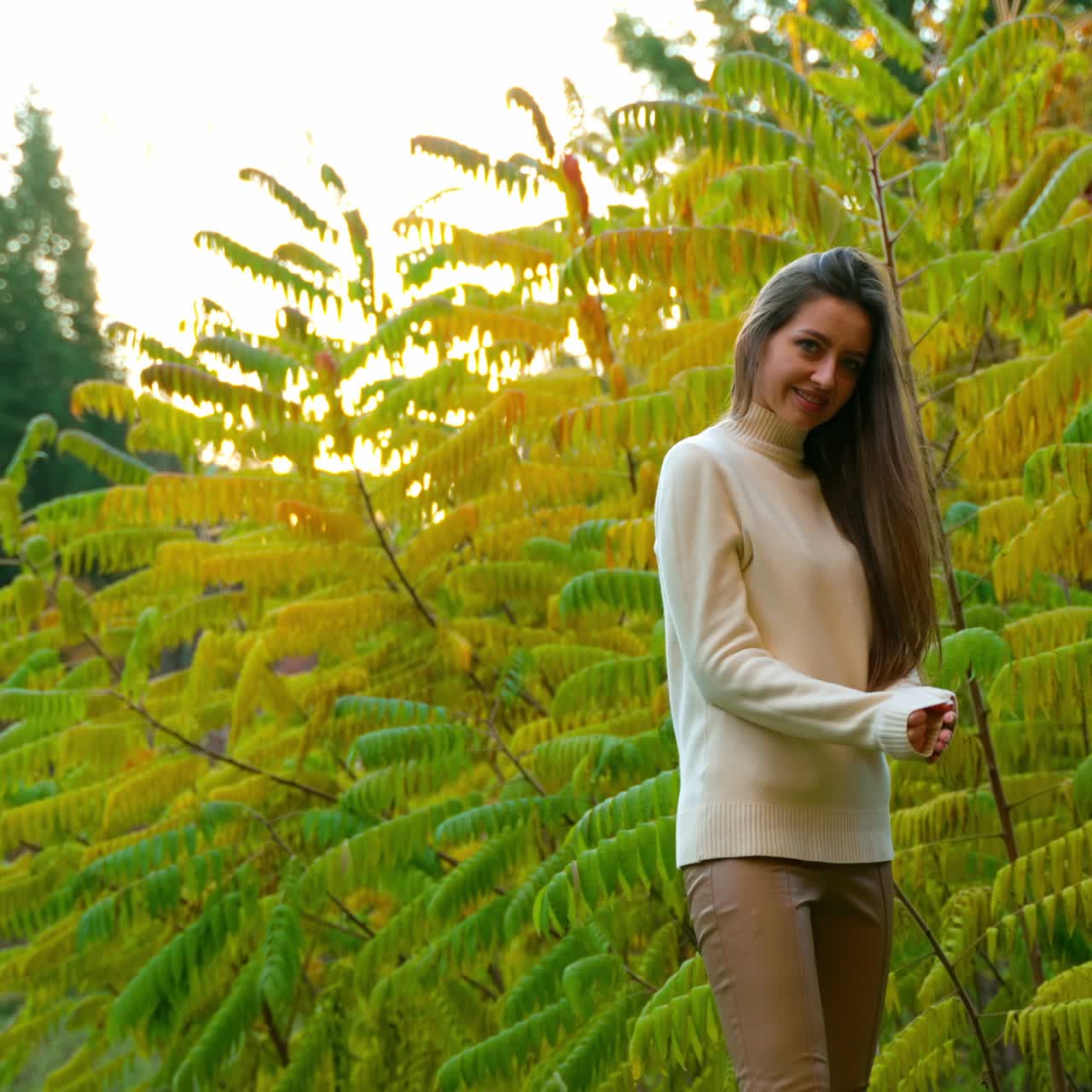 Young woman posing in front of green and yellow bush. Lady in white sweater smiling to the camera. Autumn nature at backdrop