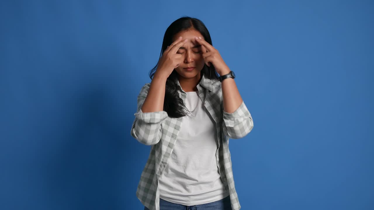 Frustrated young Asian woman with headache and frowning while standing on blue background