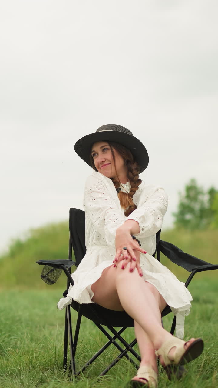 하 드레스와 검은 모자를 입은 예쁜 여자가 풀에 있는 의자에 앉아 있고, 편안하고 자신감 넘치는 태도로 포즈를 취하고 있다.