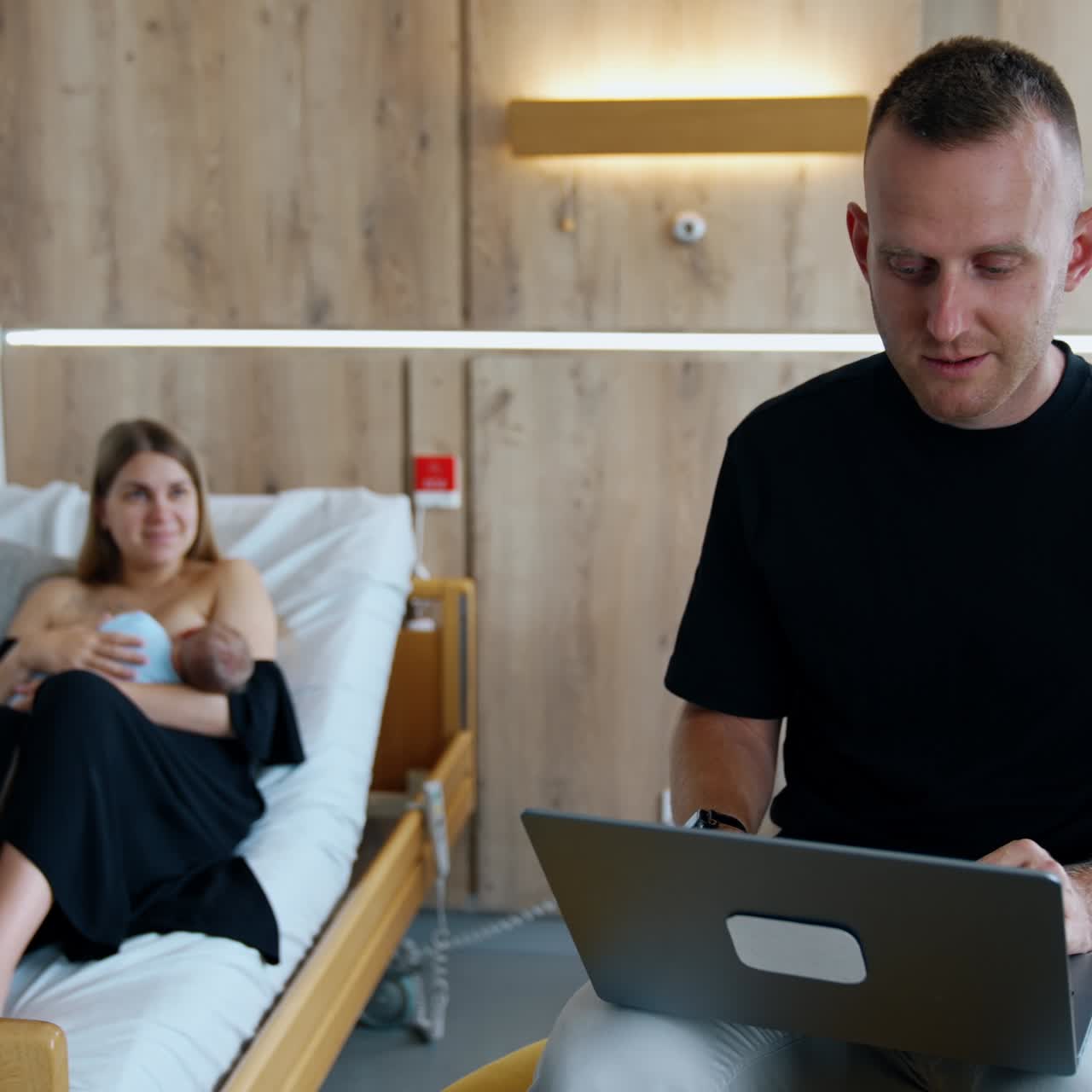 A new family of three. Parents with a newborn in a hospital. Father works at laptop while mother breastfeeding a child