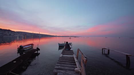 Vista-Trasera-De-Un-Joven-Refugiado-Sentado-Al-Final-Del-Muelle-De-Madera-Con-Una-Hermosa-Puesta-De-Sol-Roja-En-La-Isla-De-Lesbos,-Grecia,-Lesbos