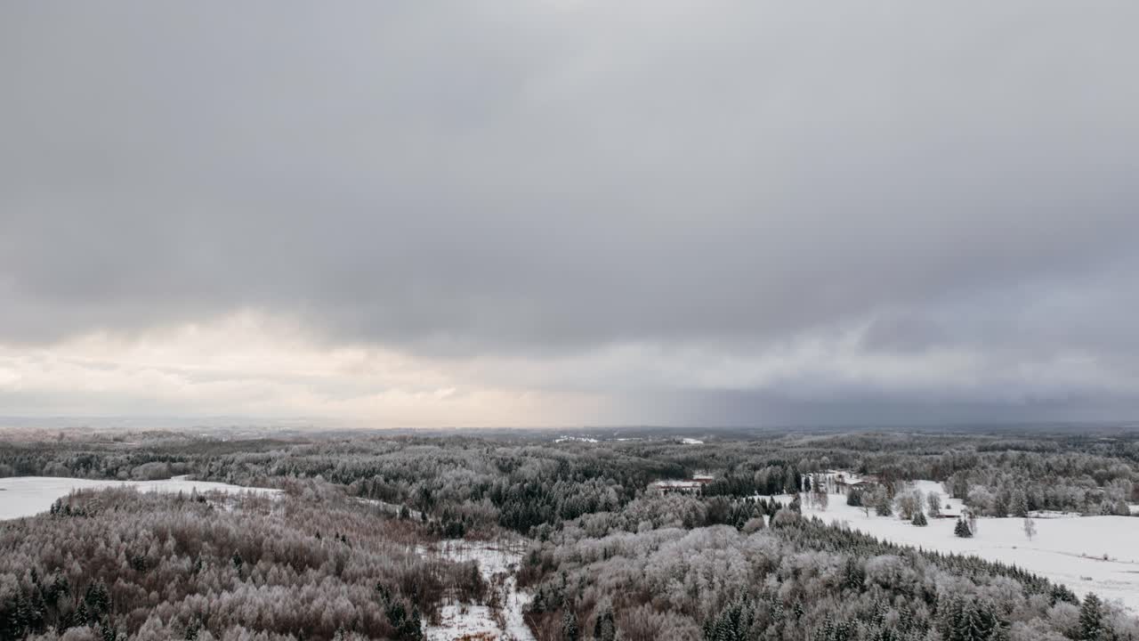 Aerial drone hyperlapse of snow covered forest and fields under dramatic winter sky with fast moving clouds in countryside.