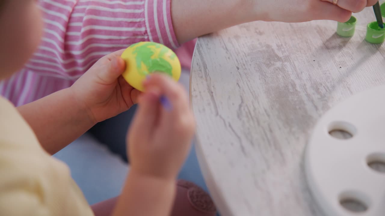 Kind bemalt kleines gelbes Spielzeug mit grünem Pinsel. Konzentrierte, sanfte Hände zeigen Kreativität. Auf einem weißen Tisch stehen bunte Farbtuben. Ideal für Bildungs-, frühkindliche Entwicklungs- oder Bastelprojekte.