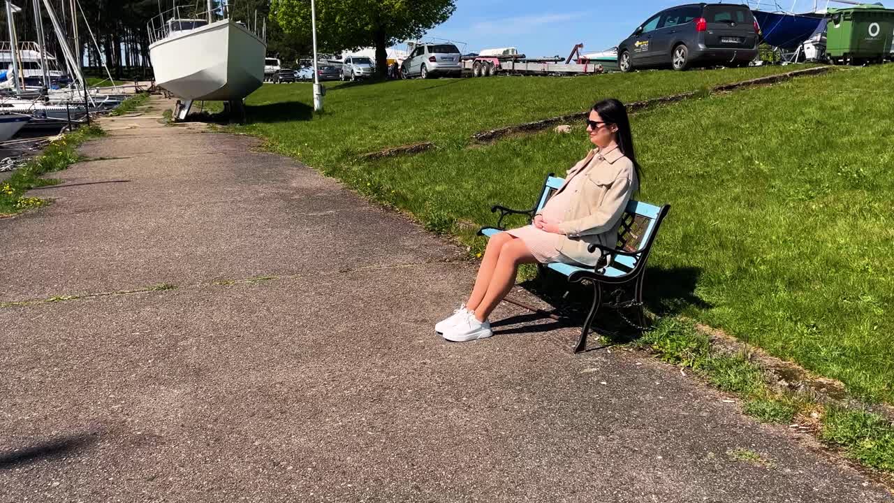 선글라스를 착용 한 젊은 임신 한 여성은  ⁇ 은 평온한 여름 날에 작은 요트의 전망과 함께 항구의 벤치에 앉아 있습니다.