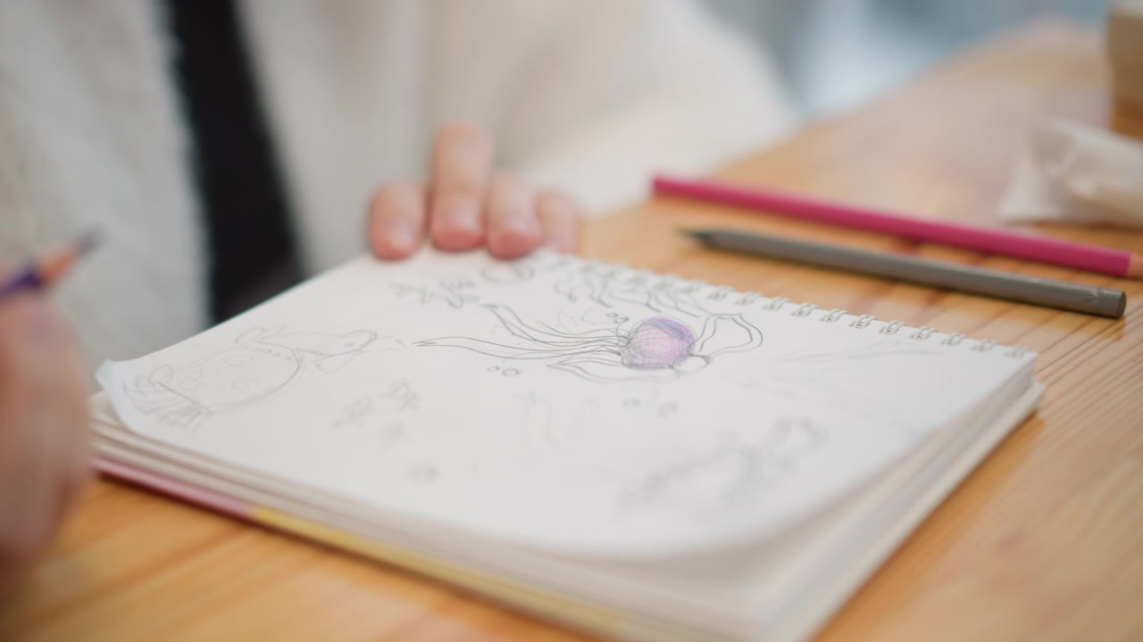木製のテーブルの上で、子供の手が鉛筆でスケッチブックに活発に絵を描き、色を塗っている。柔らかい自然光、はっきりとした跡、集中した様子、優しい動き、創造的な家庭での活動。