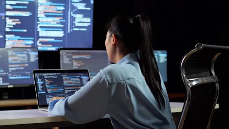 Back View Of Asian Female Programmer Leaning On Table While Writing Code By A Laptop Using Multiple Monitors Showing Database On Terminal Window Desktops In The Office