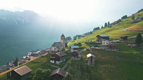 Drone-flying-backwards-from-historical-hillside-town-surrounded-by-farmland-in-the-Dolomites-Italy-with-mountain-range-in-the-background-at-golden-hour-sunset-in-summer