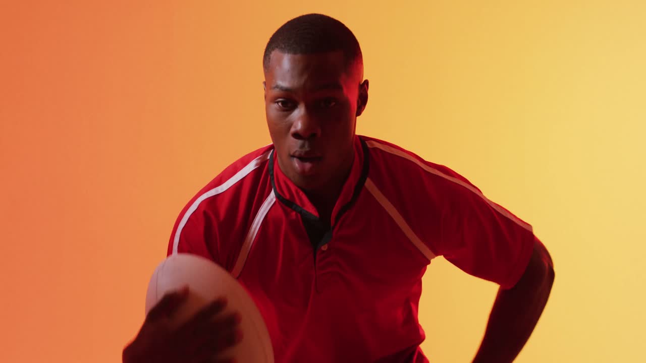 Portrait of african american male rugby player running with rugby ball over orange lighting