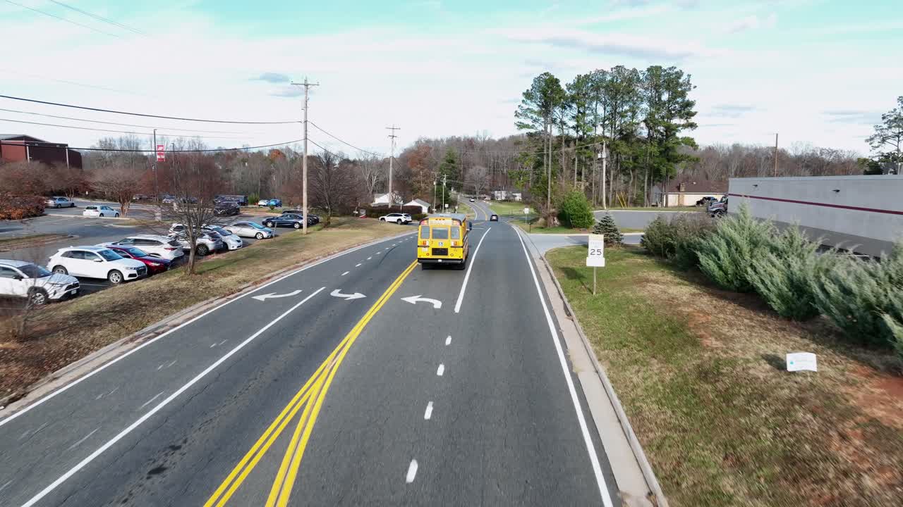 Aerial tracking of shot of yellow school bus making turn on quiet America fall roadway, surrounded by bare trees, small homes, parking lot and distant blue mountains under clear afternoon light