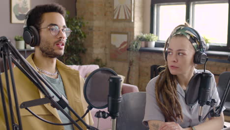 joven y mujer con auriculares sentados en una mesa con micrófonos mientras graban un podcast