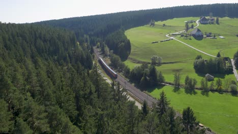 Luftaufnahme-Eines-Roten-Zuges-Der-Deutschen-Bahn,-Der-Bei-Tageslicht-Durch-üppige-Grüne-Landschaft-Mit-Dichten-Wäldern-Und-Ländlichen-Häusern-Fährt