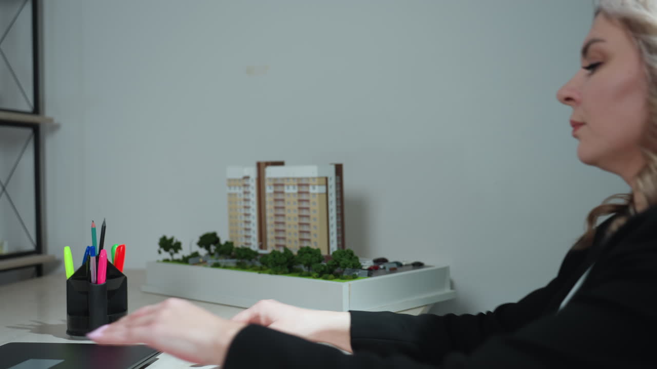 Sales woman carefully places architectural building model on desk, then sits down and pulls laptop forward to begin work, captured in stylish modern office with minimal decor and focused atmosphere