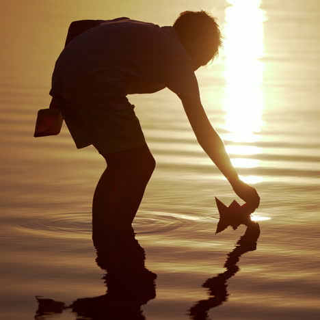 boy launches red and green paper ships in the river. origami paper boats in kid's hands. beautiful waves ripple background. travel and nature concept. video at sunset.