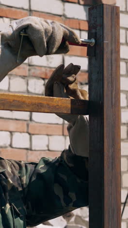 skilled builder in textile gloves installs fixing clamp on fence carcass top near brick house on sunny day