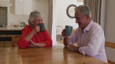 Happy-caucasian-senior-couple-sitting-at-table-in-dining-room,-smiling-and-drinking-cups-of-coffee