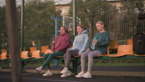 group of ladies sitting on stadium seats holding water bottles, one lady crossing her legs while another stretches her leg forward, demonstrating casual outdoor fitness session