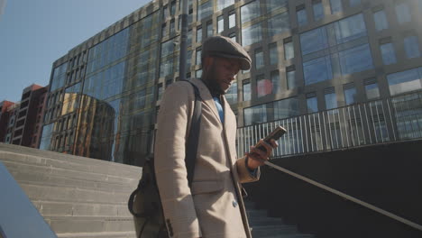 african american businessman walking in city and using smartphone