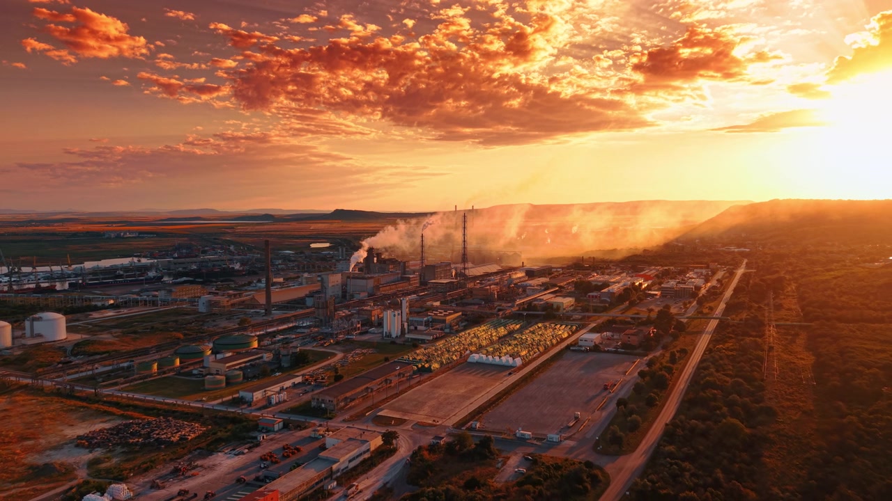 Complexo industrial ao pôr do sol com nuvens laranja brilhantes. Fumaça sobe das chaminés sobre estruturas de fabricação ao lado de uma estrada reta sob os raios do sol.