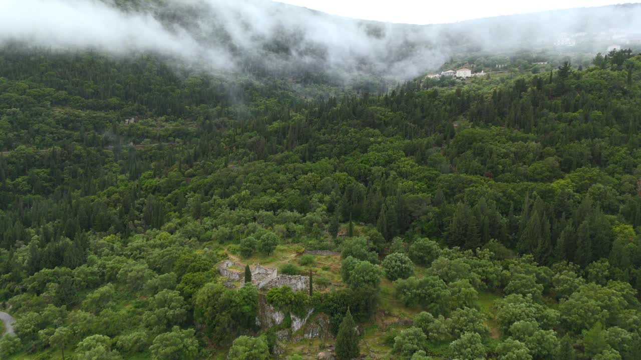 Aerial drone view of the ancient ruins believed to be the Palace of Odysseus on Ithaca Island, Greece with cloudy mountain summits and green forests surroundings