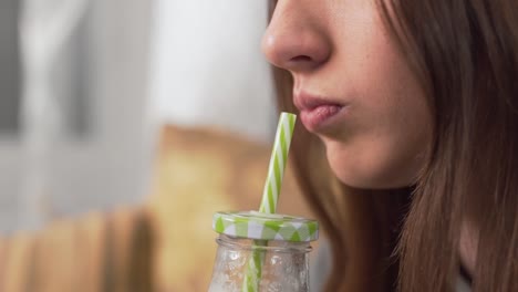 escena de primer plano de una hermosa chica que está sentada en casa en el sofá, bebiendo un batido casero de una botella con una pajita