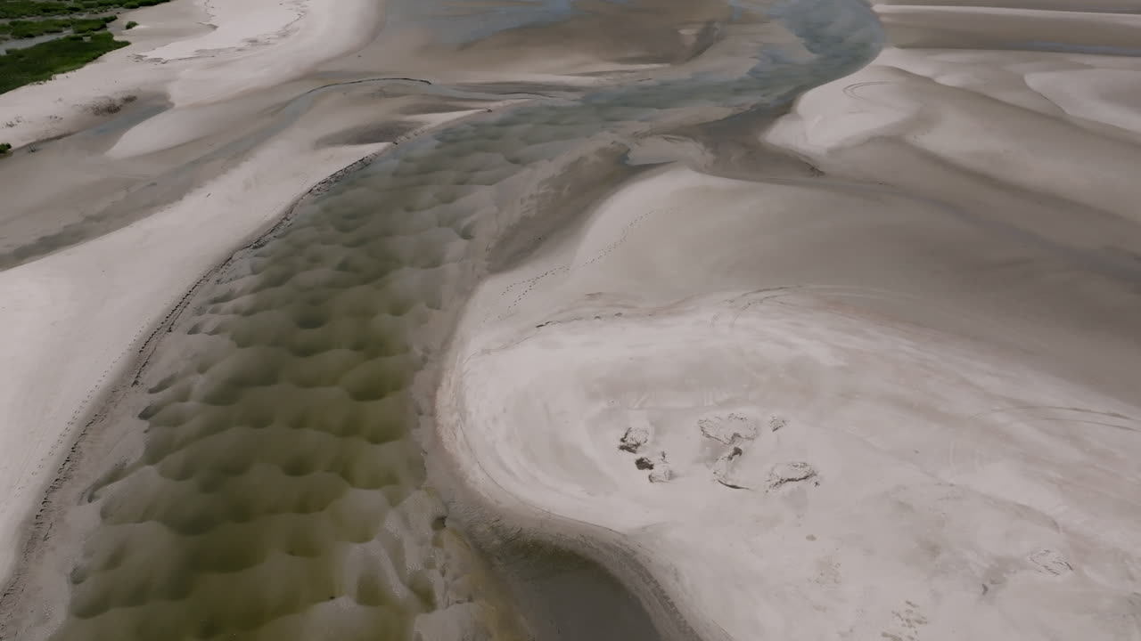 Un ruscello di marea sinuoso si snoda attraverso la sabbia modellata, dove i segni dei pneumatici e un camion rosso offrono un senso di scala rispetto alle vaste forme scolpite della natura.