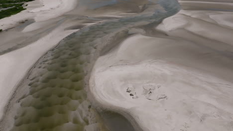 a snaking tidal creek cuts through textured sand, where tire marks and a red truck offer scale against nature’s vast sculpted forms