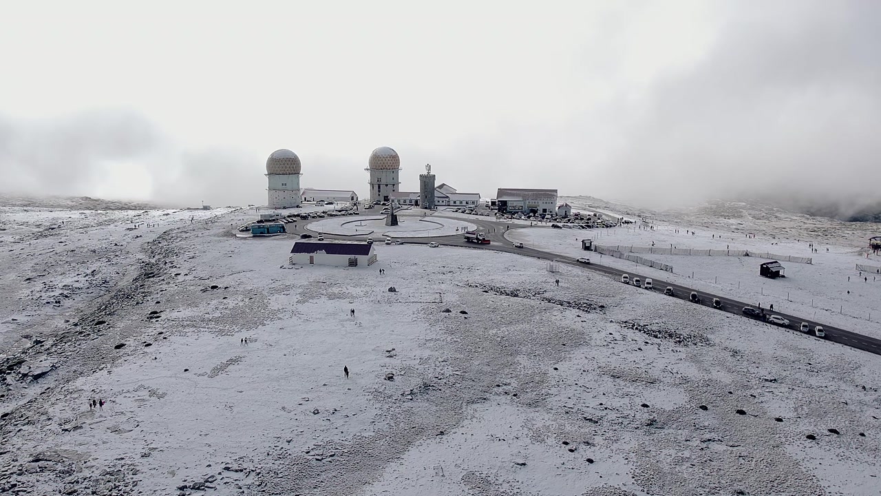 Drone view of Torre, highest point of Serra da Estrela, Portugal, at N339 road, showing radar domes, summit buildings, parked vehicles and snow-covered plateau, aerial orbit
