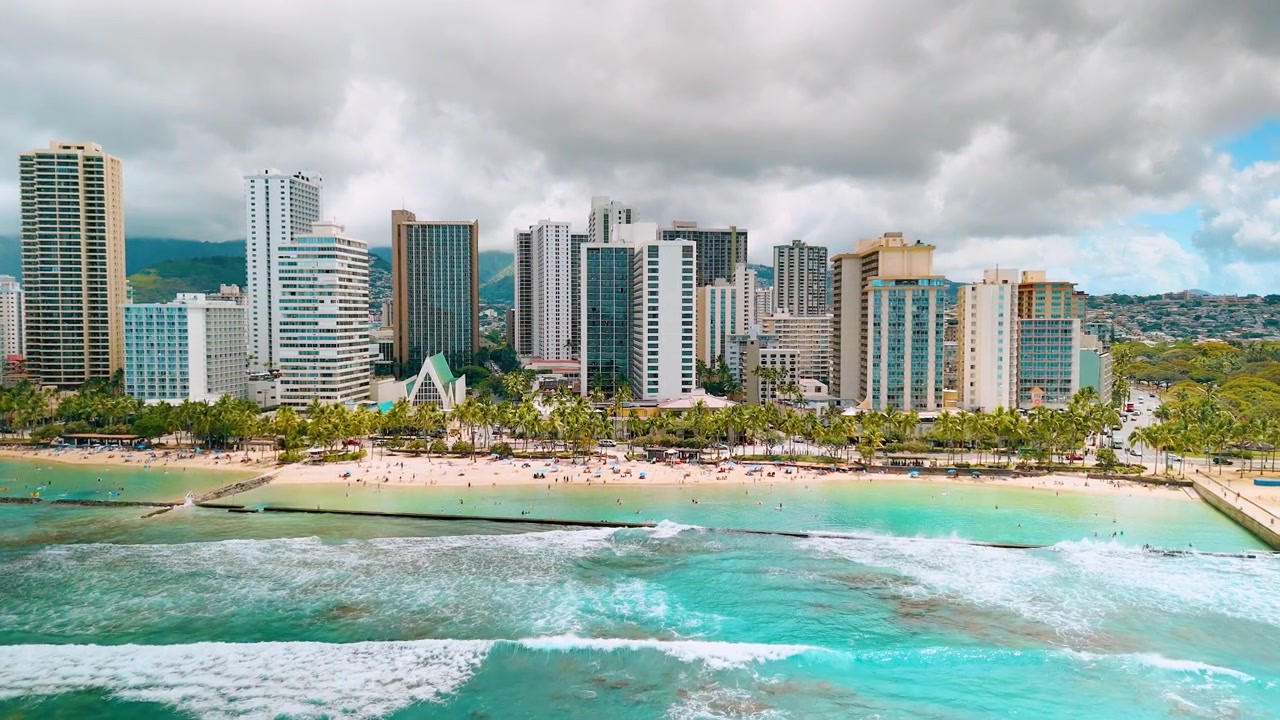 Cielo coperto sopra le torri della spiaggia di Waikiki, Honolulu, Hawaii. Onda turchese che si infrange vicino alla riva sabbiosa, torre dell'hotel e palma