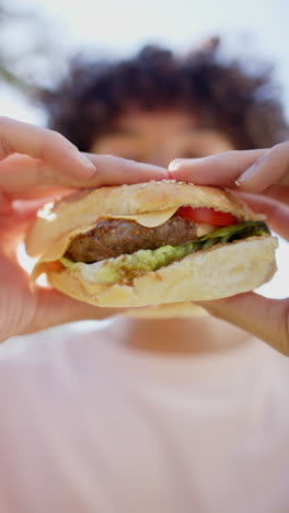 vertical video: holding juicy burger outdoors, person savoring sandwich with fresh ingredients