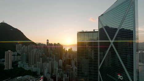 Toma-Aérea-Reveladora-De-Una-Puesta-De-Sol-Dorada-Detrás-Del-Banco-De-China-Y-Un-Denso-Desarrollo-Urbano-En-La-Ciudad-De-Hong-Kong