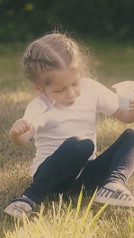 nice little girl with fair hair eats fresh yogurt with spoon sitting on grass at green sunny park slow motion