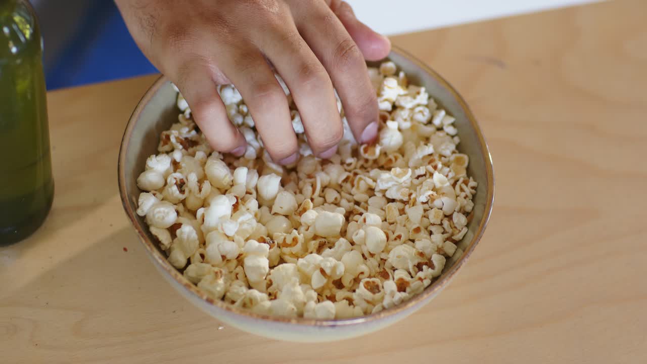 Uomo afroamericano di mezza età che allunga la mano in una ciotola, prendendo popcorn, facendo uno spuntino al tavolo con una bottiglia.