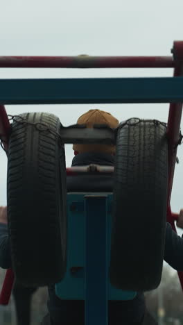 back view of a coach exercising on outdoor equipment, lifting weights with red bars as stops and moves his body and lifts his right leg, while two boys are working out on a track