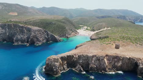 Boat-Sails-out-Blue-Cove-at-Cala-Domestica-Beach,-Italy---4k-Aerial-Circling